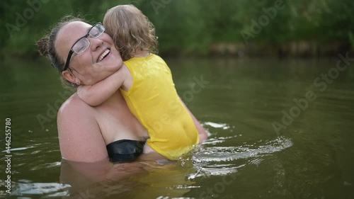 Little funny cute blonde girl child toddler in yellow bodysuit laughing learns swim outside at summer lake. Plus size body positive smiling woman mother baby swimming in natural pool. Water splashes