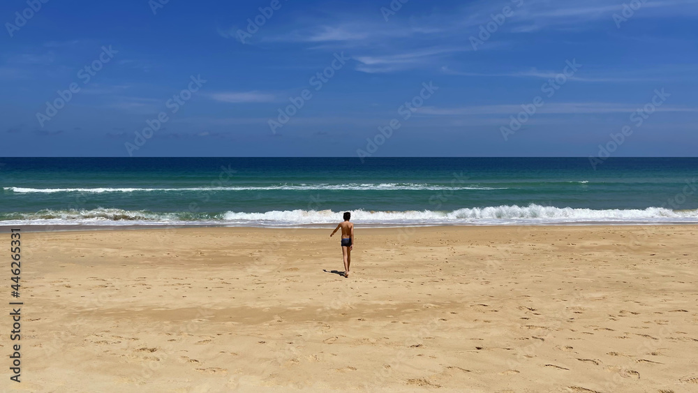 Wide panorama. Seascape. Blue sky, open ocean, yellow sand. One boy goes towards sea. Beautiful view of tropical beach. Small waves. Line of horizon. Expanse of turquoise water. Boy is in the center 