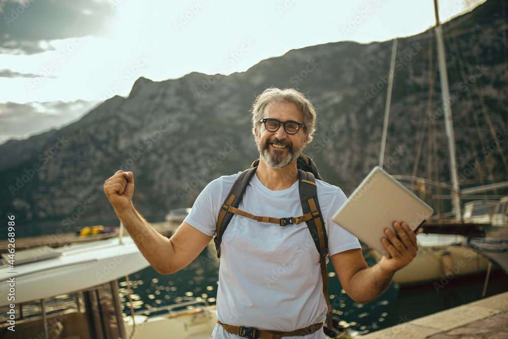 Smiling tourist mature man standing with digital tablet and backpack near the sea. Smiling tourist mature man standing with digital tablet and backpack near the sea.