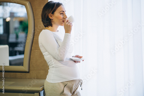 Young pregnant woman standing by the window and drinkig coffee photo