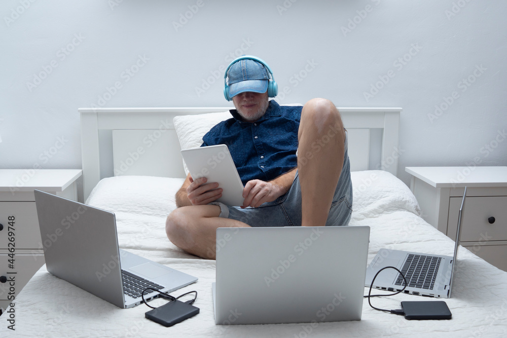 man sitting on bed with headphones with several laptops and peripherals man sitting on bed with headphones with several laptops and peripherals