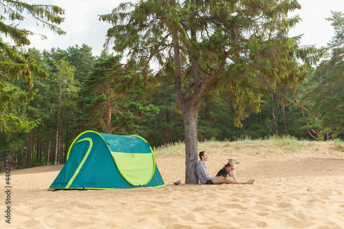 Man with Dog by tent under a tree on a sandy beach