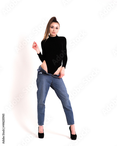 The brunette stands on a white background in a black bodysuit, jeans and high heels