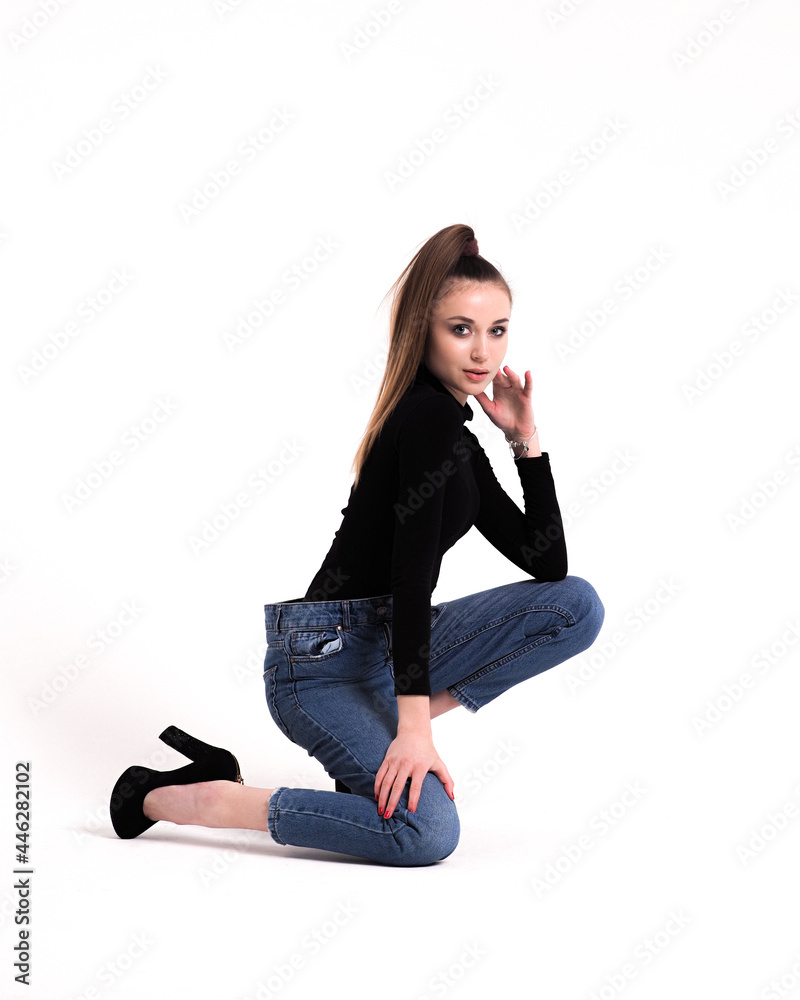 The brunette is on her knees on a white background in a bodysuit and jeans with heels and a ponytail The brunette is on her knees on a white background in a bodysuit and jeans with heels and a ponytail