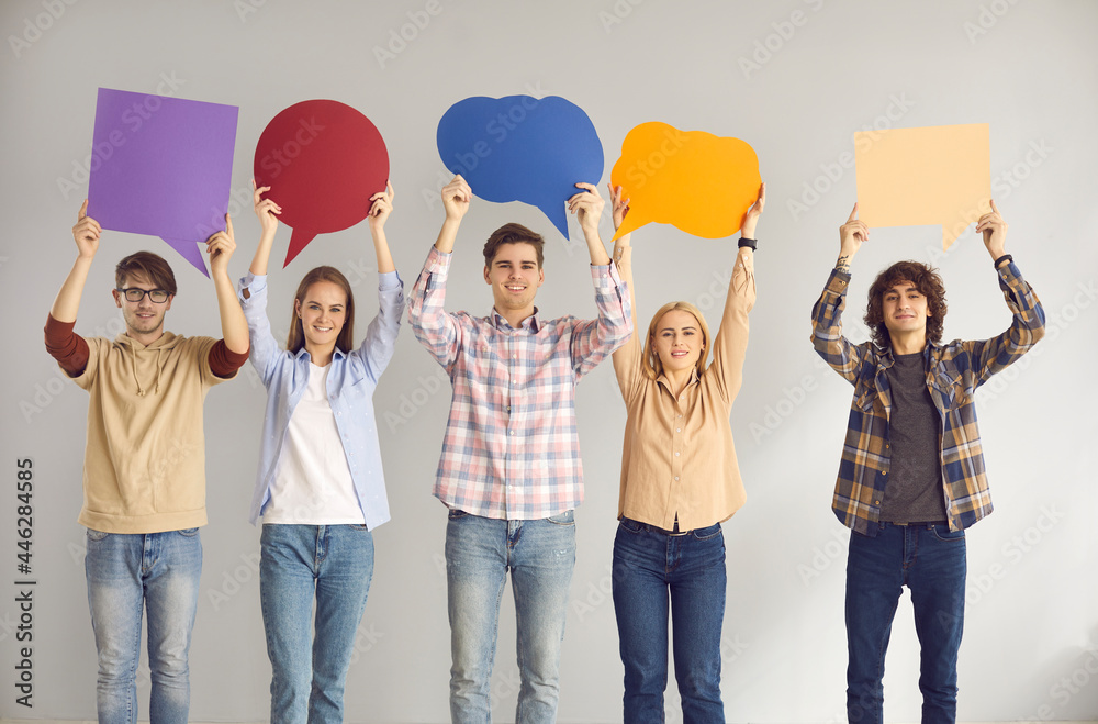 Naklejka premium Group of happy smiling young people holding up multi colored cardboard and paper mockup speech bubbles standing in studio with gray background. Expressing opinion and sharing important message concept