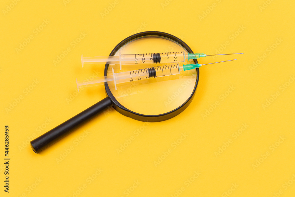 A Magnifier with Medical Syringe Lying on Yellow Table - Vaccination Concept A Magnifier with Medical Syringe Lying on Yellow Table - Vaccination Concept