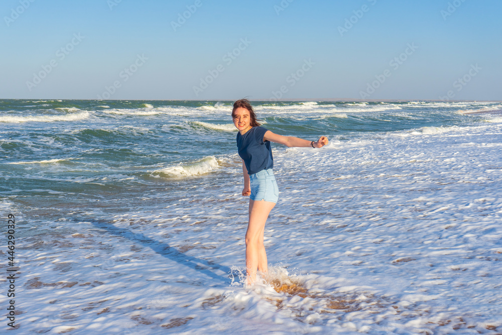 Tween slender girl standing on sand beach of sea coast against stormy waves in sunny summer day. Young woman spending summer weekend at sea. Trip to seaside, summer vacation concept