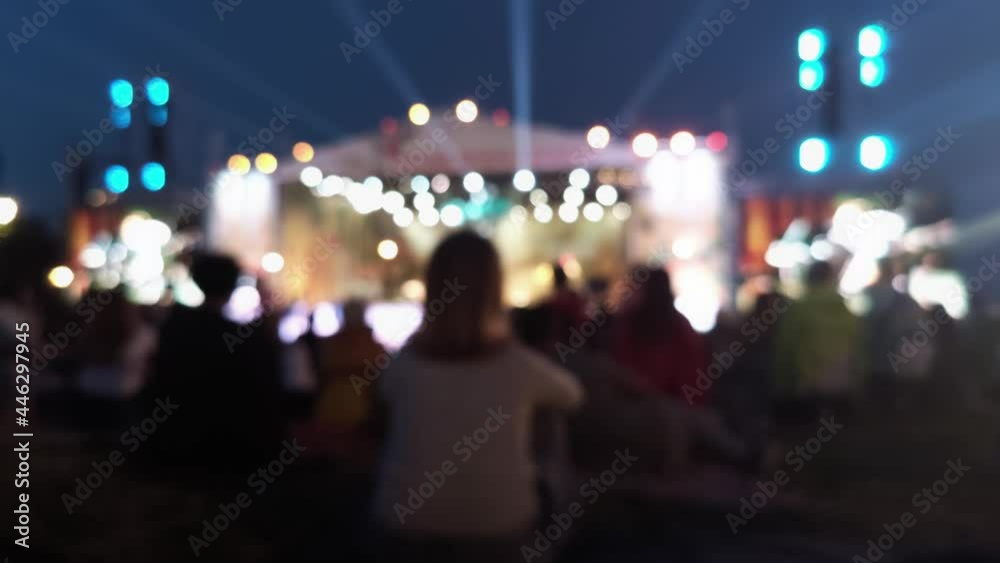 Girl sitting on a green field at a festival. Blurred image of people in a festival. Night lights Girl sitting on a green field at a festival. Blurred image of people in a festival. Night lights