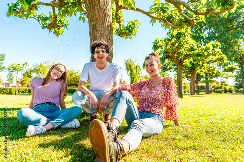 Three young diverse friends resting on green park field smiling looking at camera for a portrait. Gen z students spending time in nature to breaking city life routine. Friendship makes no difference