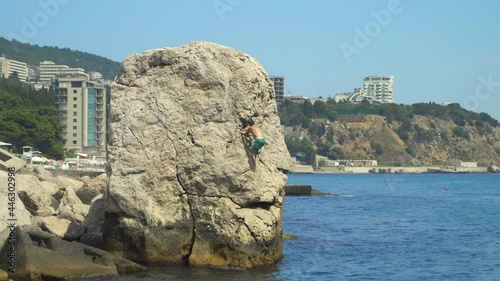 A young man climbs a high rock in the sea to jump into the water on a hot summer day.