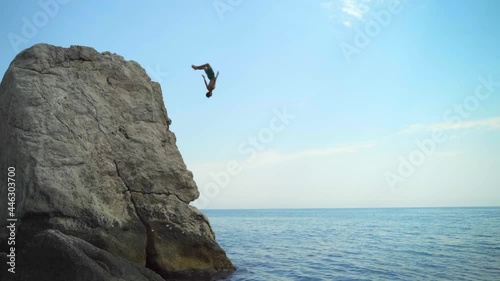 A young man jumping from a high cliff into the sea doing a back somersault in slow motion on a hot summer day