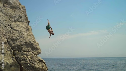 A young man jumping from a high cliff into the sea doing a back somersault in slow motion on a hot summer day