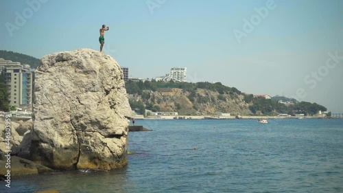 A young man jumps from a high cliff into the sea making beautiful turns in the air and enters the water with his feet in slow motion on a hot summer day