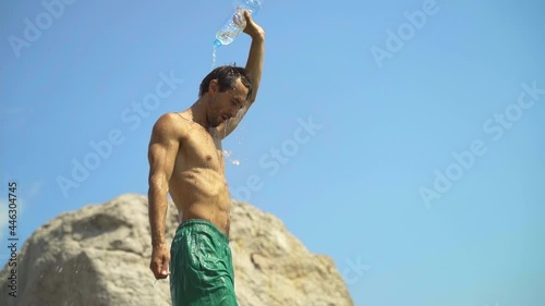 A young guy of athletic build in the heat is poured with fresh water from a plastic bottle. The guy is doused with water from the heat in slow motion and smiles contentedly.