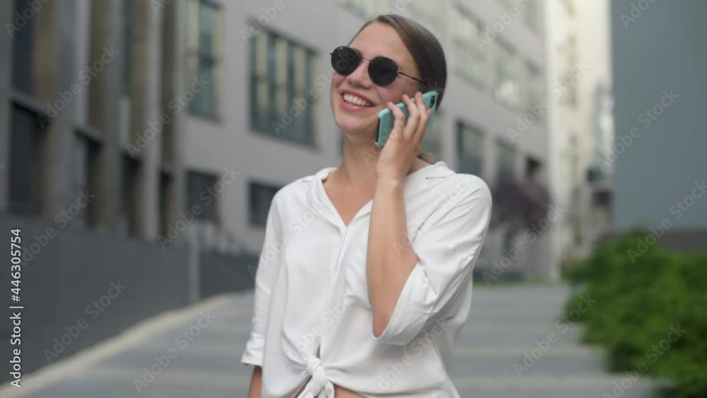 Smiling lady holding cellphone in hands outside. Business woman wearing trendy sunglasses walks down the central city street and uses her phone. Smiling lady holding cellphone in hands outside. Business woman wearing trendy sunglasses walks down the central city street and uses her phone.