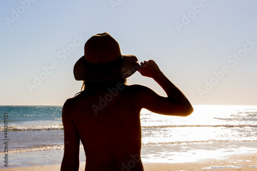 Young girl holding hat looking at the sea at sunset. Selective focus. Backlighting.