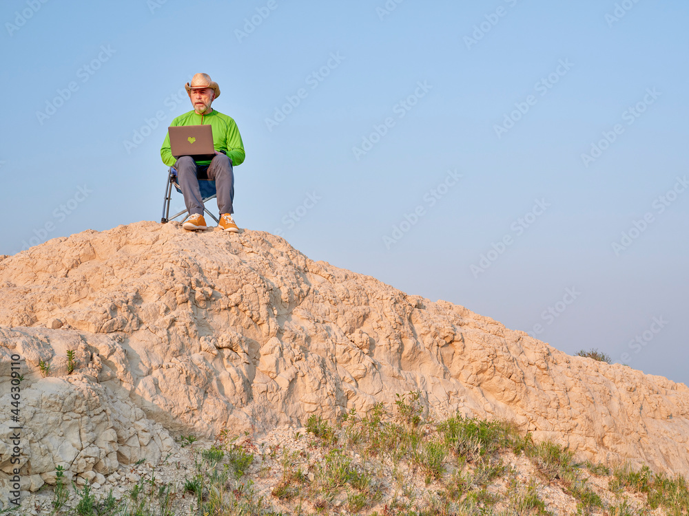 Fototapeta premium Senior man in cowboy hat is sitting on a folding chair and working on laptop in the middle of nowhere, early morning in the badlands of Pawnee National Grassland in Colorado