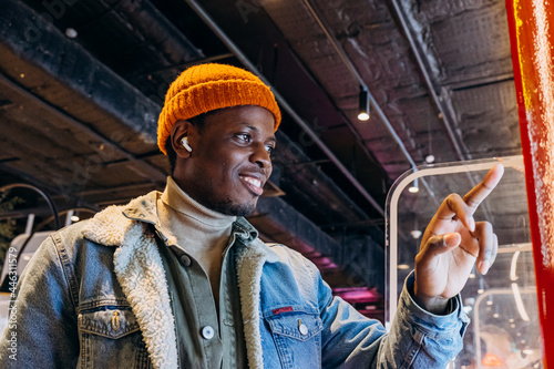 Obraz na plátně Smiling African-American man in warm denim jacket with wireless earphones uses s