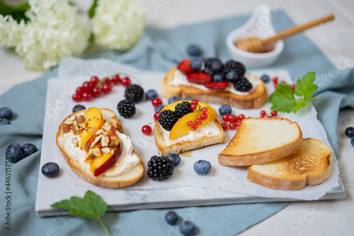 Toasted bread with cream cheese, honey, nuts, fruits and fresh berries. Summer tasty breakfast - dessert on white textured table. Selective focus with copy space.