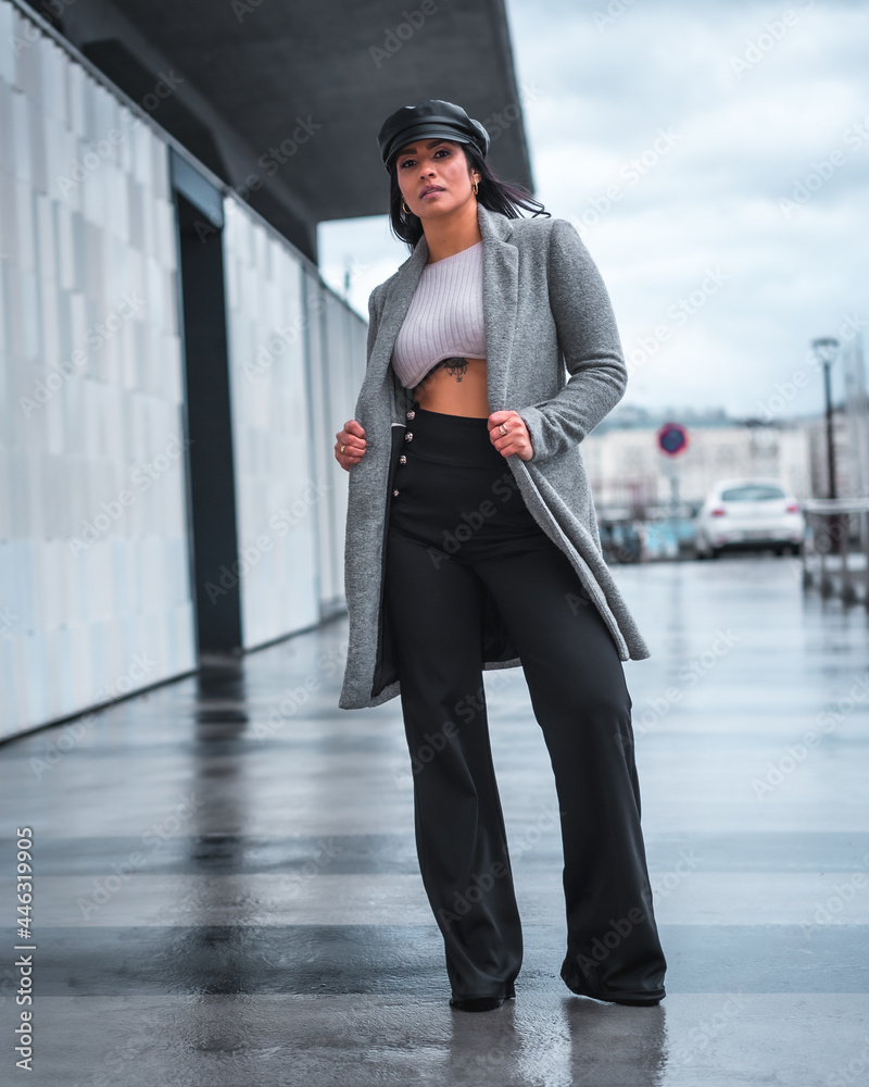 Obraz premium Street style in the city of a beautiful Latin girl with a leather hat and a gray raincoat walking in autumn