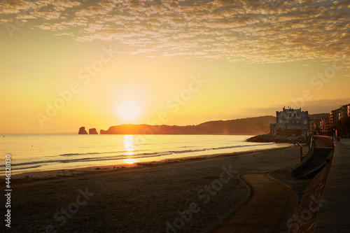 sunrise on the beach, a close-up of the sand and in the background two large twin rocks next to the cliff, a couple walking near an old building © arakawa
