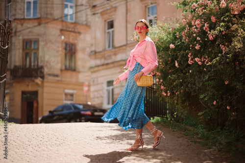 Summer street fashion conception: elegant woman wearing trendy pink sunglasses, shirt with knot, polka dot blue midi skirt, white strap sandals, with yellow shoulder bag, walking in street. Copy space photo