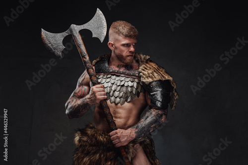 Brutal tattooed warrior wearing light armour and fur holding two-handed axe in dark studio © Fxquadro