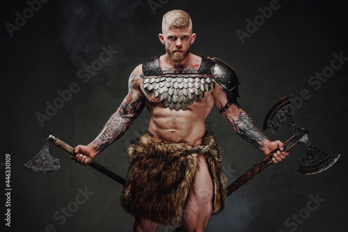 Photo Brutal tattooed warrior wearing light armour and fur holding axes in dark studio