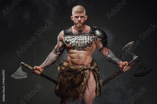 Fotografie, Obraz Strong tattooed warrior in light armour and fur with two axes in dark