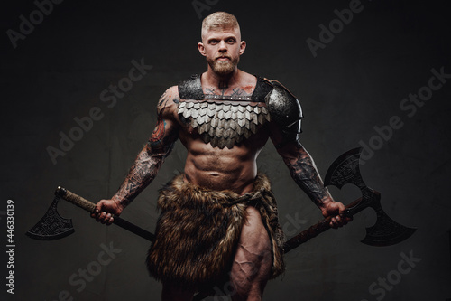 Brutal tattooed warrior wearing light armour and fur holding axes in dark studio