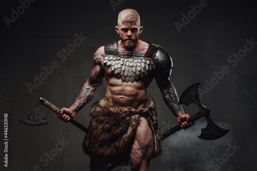 Strong tattooed warrior in light armour and fur with two axes in dark