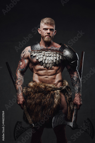 Brutal tattooed warrior wearing light armour and fur holding axes in dark studio