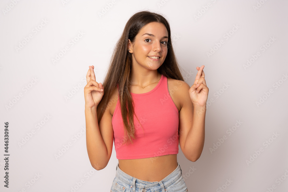 Portrait of a smiling young girl with long brunette hair standing over white background, holding fingers crossed for good luck Portrait of a smiling young girl with long brunette hair standing over white background, holding fingers crossed for good luck