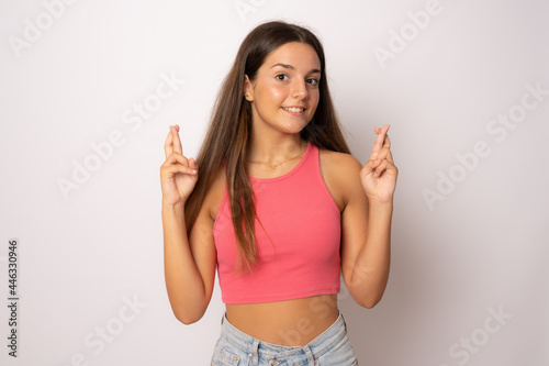 Portrait of a smiling young girl with long brunette hair standing over white background, holding fingers crossed for good luck