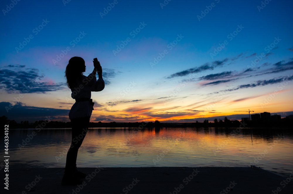 Silhouette of a girl with a phone on the background of sunset by the lake