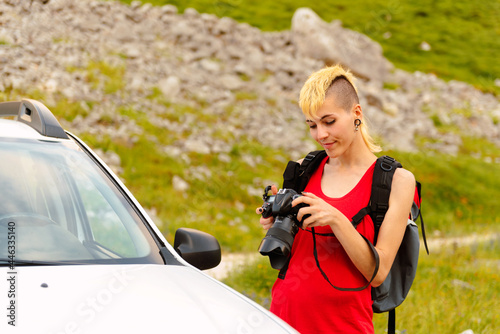 Young Caucasian girl with backpack near her off-road car holding a photo camera and reviewing the pictures taken during a nature trip. © Alberto