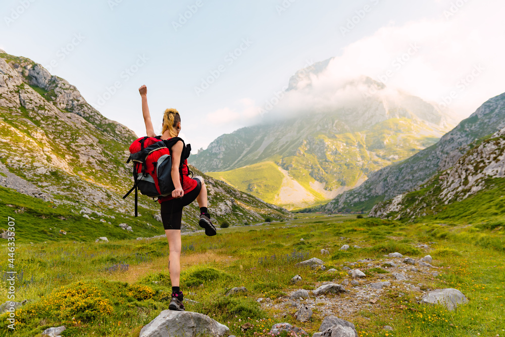 Caucasian and young person with his back turned, with a backpack and on top of a rock lifting one leg and one arm. Happy white woman hiking up the mountain