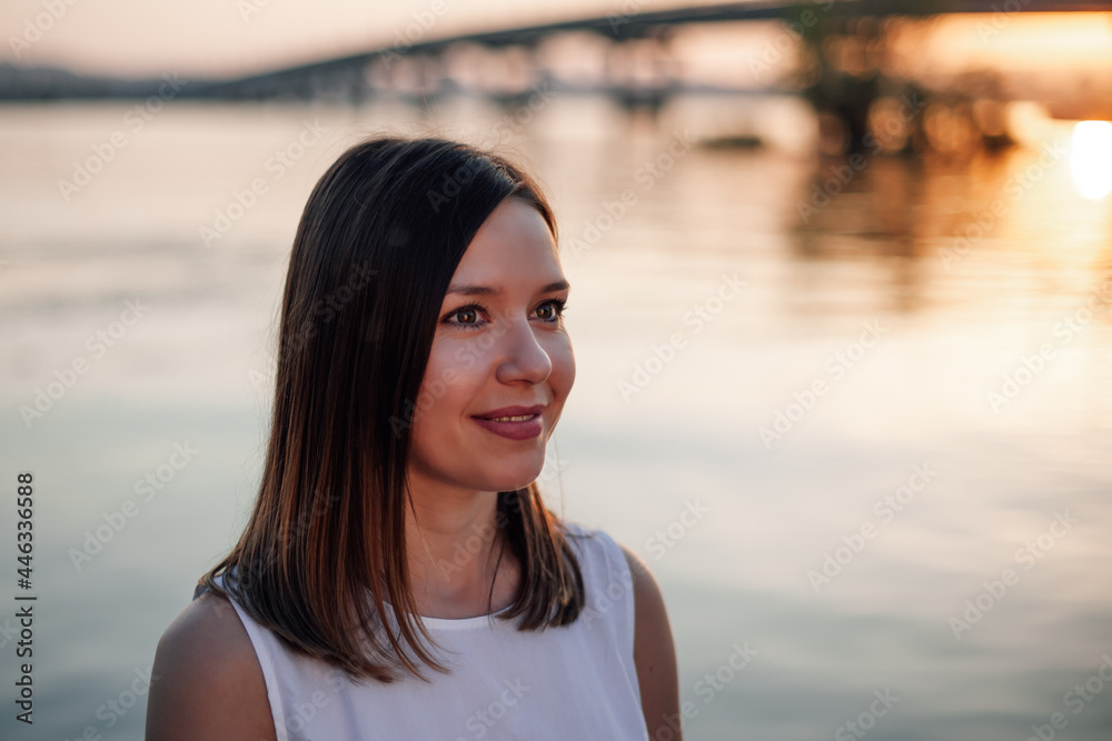 Portrait of a woman at sunset. Close-up portrait of a charming Caucasian woman looking at the sunset on a summer evening at the bridge over the river. 