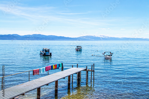 Dock with beach towels drying  stretching out onto Tahoe lake with boats moored offshore and snowtopped mountains and a pastel sky in the distance - Beautifully turqoise photo
