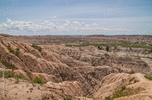 Badlands National Park, SD, USA - June 1, 2008: Landscape of beige-brown canyons and walls of geological deposits under light blue cloudscape Green patches and singular trees. photo