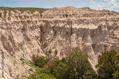 Badlands National Park, SD, USA - June 1, 2008: Looking into canyon with its beige geological deposit flanks. Green trees down and light blue sky above.