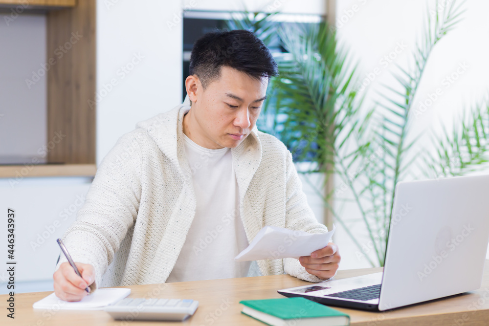 Fototapeta premium Young Asian man doing paperwork at home sitting at a desk, working on a laptop, focused on paying bills
