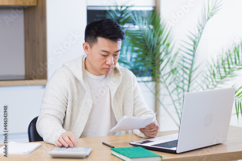 Young Asian man doing paperwork at home sitting at a desk, working on a laptop, focused on paying bills