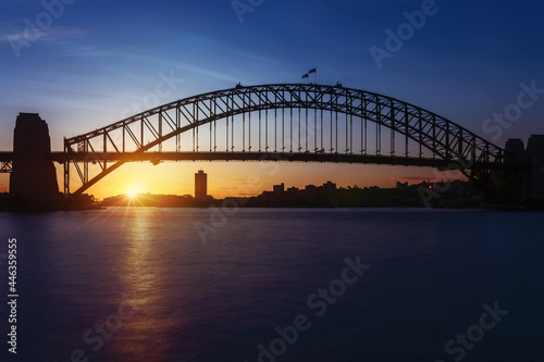 Sun setting behind the iconic Sydney Harbour Bridge in Sydney Harbour late in the evening in Australia with the silhouette of the city of North Sydney in the background.