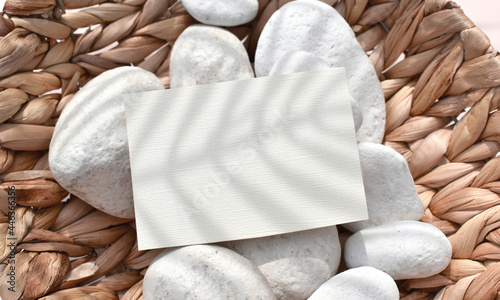 Close up empty paper blank, invitation card on pebbles in rattan basket. Layout summer still life concept for wedding invitation, vacation or spa branding. Sunlit eco mockup scene.
