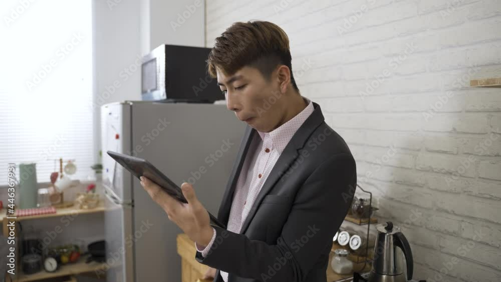 portrait of an asian business man reading online news on an electric pad while eating bread for breakfast in the morning at home kitchen.