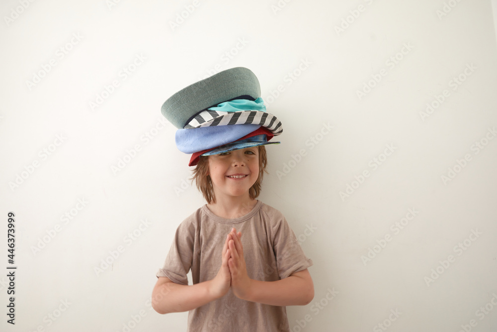 Smiling boy wearing t shirt and various headwear standing against white background and looking at camera. Child's palms are folded at the chest in gratitude