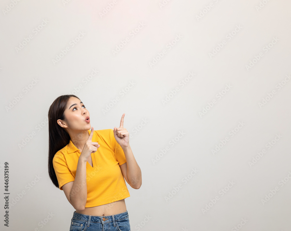 Excited asian woman pointing the finger to blank space over head for advertising text on isolated background. Joyful teenage girl in yellow shirt standing in white room looking at empty space. Excited asian woman pointing the finger to blank space over head for advertising text on isolated background. Joyful teenage girl in yellow shirt standing in white room looking at empty space.