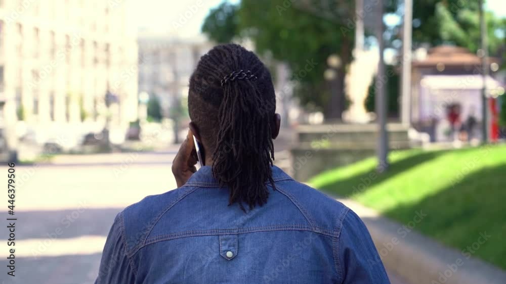Back view of African hipster man with stylish hairstyle walking on the street talking on mobile phone, slow motion Back view of African hipster man with stylish hairstyle walking on the street talking on mobile phone, slow motion