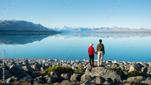 Couple standing on the shore of Lake Pukaki, holding hands and watching Mt Cook reflected in the clear waters, South Island, New Zealand